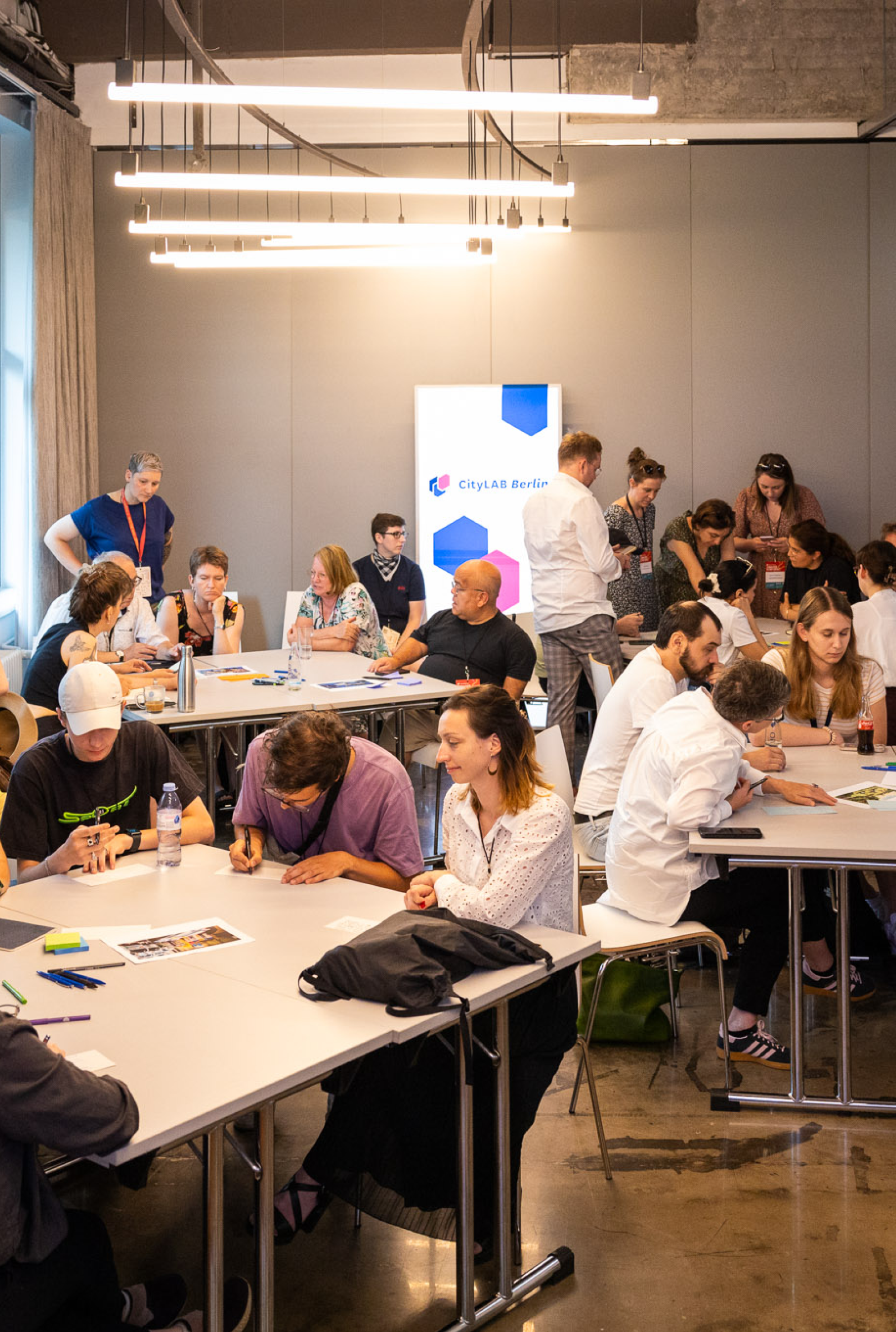 Ein Workshop raum bei der <em>CityLAB Sommerkonferenz 2024</em>. Viele Menschen sitzen an verschiedene Tische und arbeiten gemeinsamen, schreiben Notizen auf, usw. Im Hintergrund is ein CityLAB Aufsteller zu sehen.