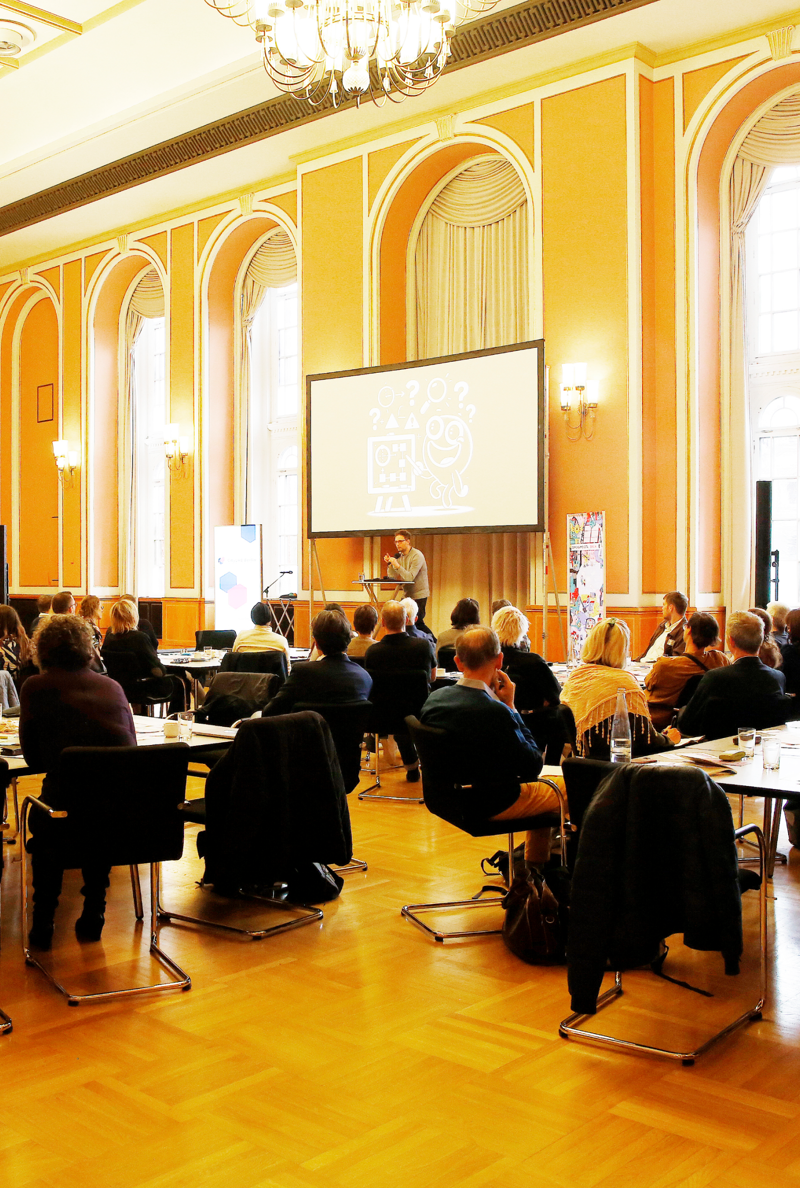 Eine Präsentation in einem eleganten Veranstaltungssaal mit gelben Wänden und klassischen Architekturdetails wie Wandleuchten und Kronleuchtern. Ein Redner steht vor einer großen Projektionsfläche, während das Publikum an Tischen im Raum sitzt. Der historische Charakter des Raums mit seinen hohen Bogenfenstern und Stuckarbeiten kontrastiert mit der modernen Präsentationstechnik und schafft eine eindrucksvolle Atmosphäre für die Veranstaltung.