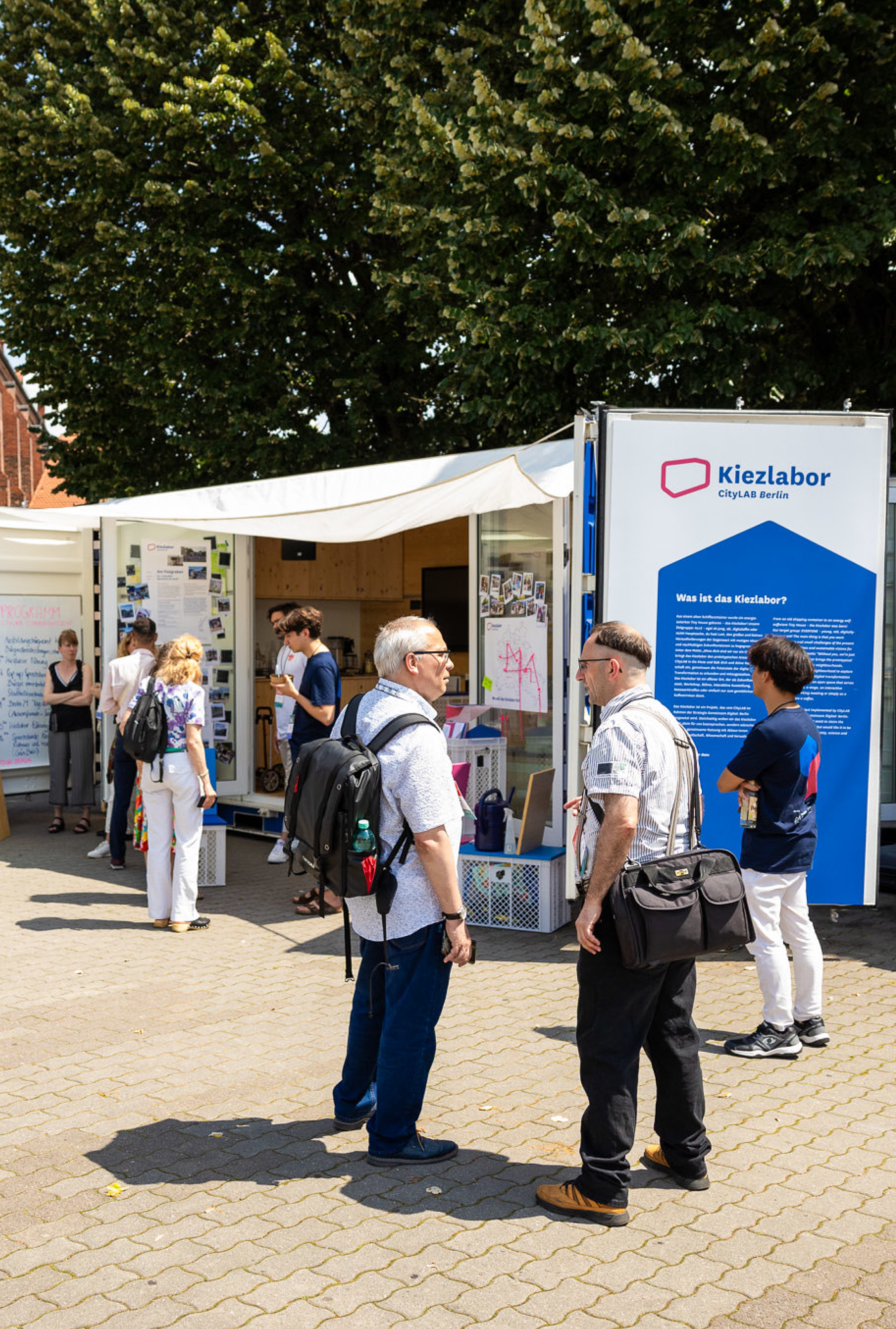 Das mobile Kiezlabor des CityLAB Berlin im Einsatz: Ein weißer Container mit Sonnensegel steht auf einem gepflasterten Platz. Mehrere Besucher:innen betrachten die Informationstafeln und Ausstellungsmaterialien. An der Außenseite ist ein großes blaues Banner mit dem Kiezlabor-Logo und Erklärungstext zu sehen. Die Szene spielt sich an einem sonnigen Tag unter einem großen Baum ab.